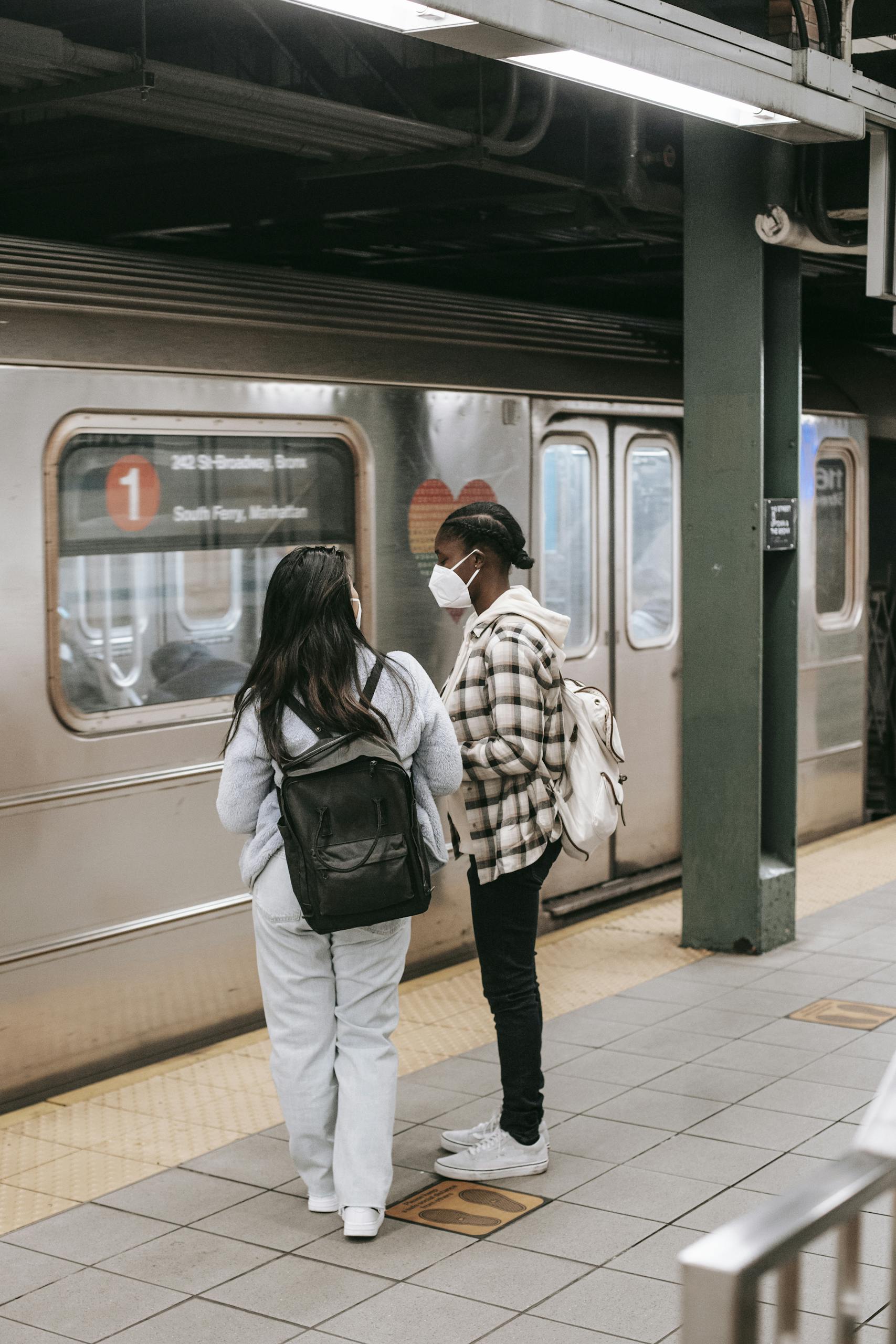 Back view of anonymous multiracial girlfriends in casual clothes with backpacks in masks against public transport in subway station