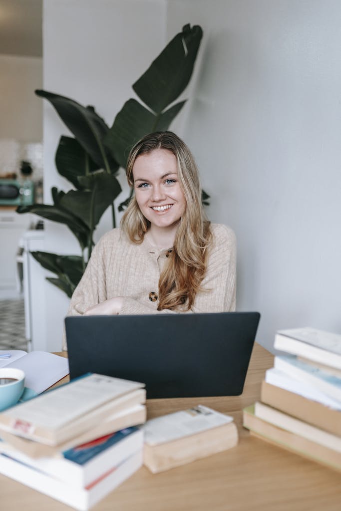 Positive young female with long hair smiling and looking at camera at table with many books and netbook